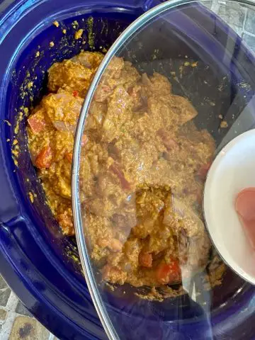 Raw chicken pieces coated with a yogurt based seasoning paste and tomatoes in a blue slow cooker with a glass lid poised over the slow cooker.
