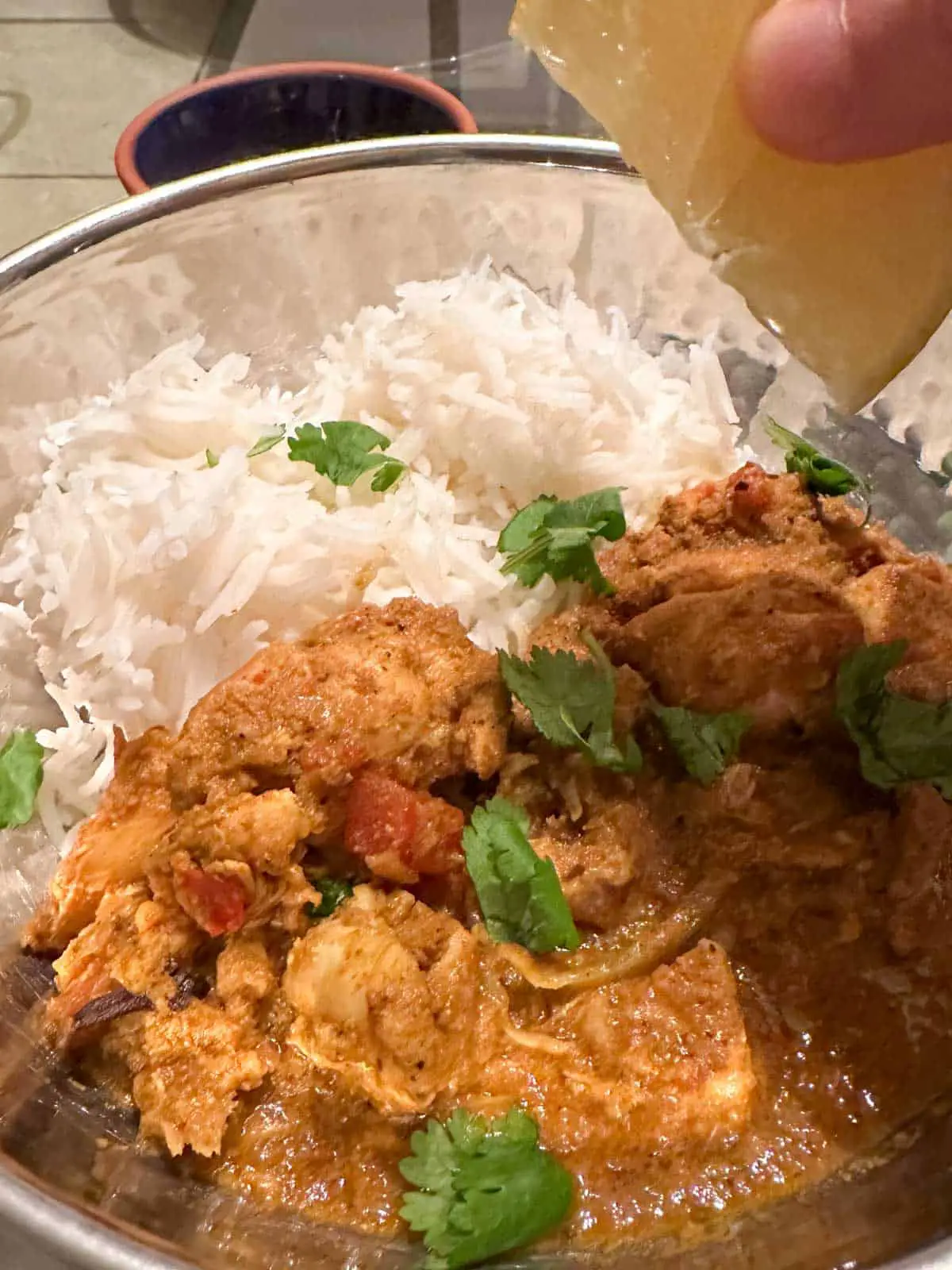 Spicy Indian Chicken Curry with basmati rice garnished with cilantro in a silver balti dish with a wedge of lemon poised over the chicken. There is a small blue bowl behind the balti dish.
