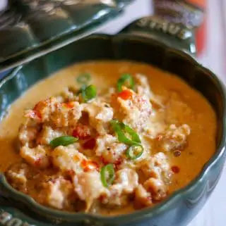 A creamy sauce containing crawfish tails in a green Staub cocotte, garnished with green onions and hot sauce. There is a bottle of Tabasco in the background.