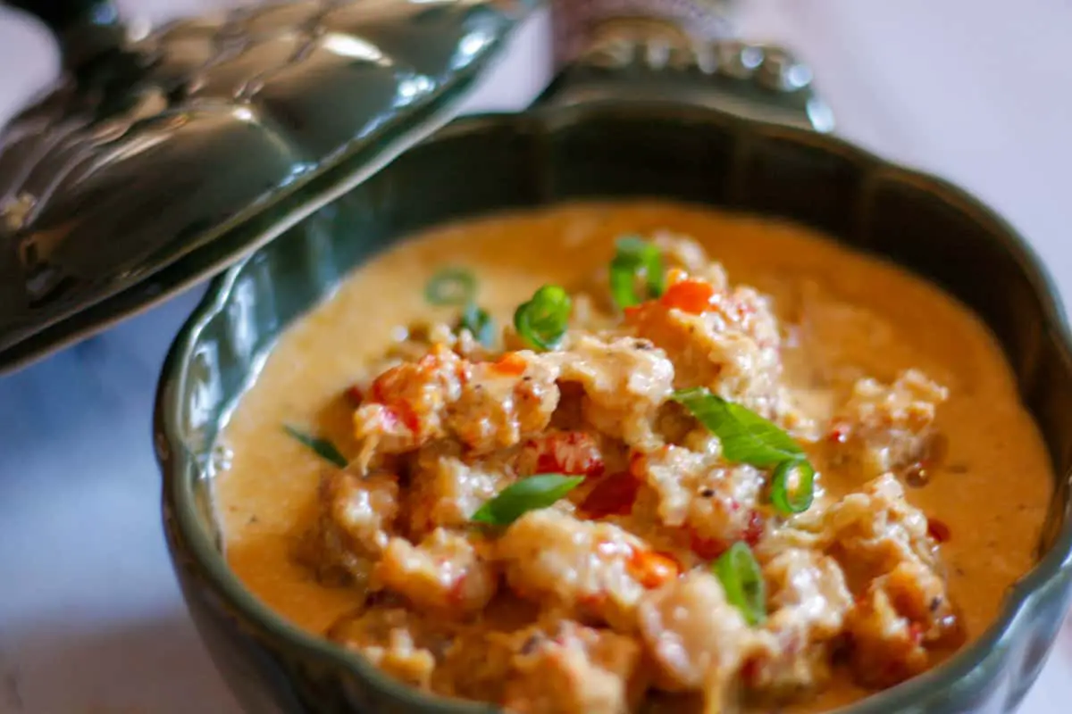 A creamy sauce containing crawfish tails in a green Staub cocotte, garnished with green onions and hot sauce.