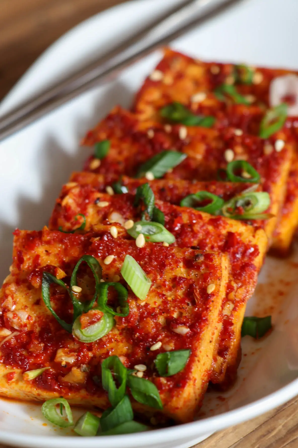 Slices of tofu coated in a spicy sauce garnished with green onions and sesame seeds in a white dish with silver chopsticks resting on the white dish.