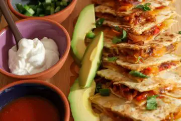 Quesadillas filled with melted cheese and sautéed vegetable mixture. There are slices of avocado and cilantro leaves garnishing the quesadilla wedges and small bowls in the background filled with salsa, sour cream, and cilantro.
