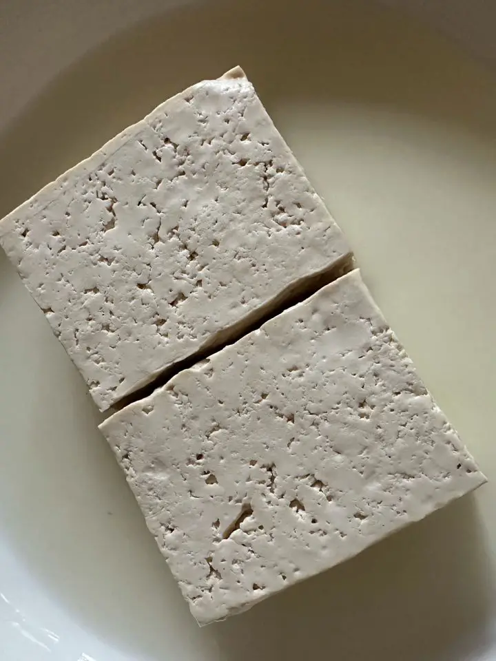 Tofu blocks sitting in drained water from the tofu in a white dish.