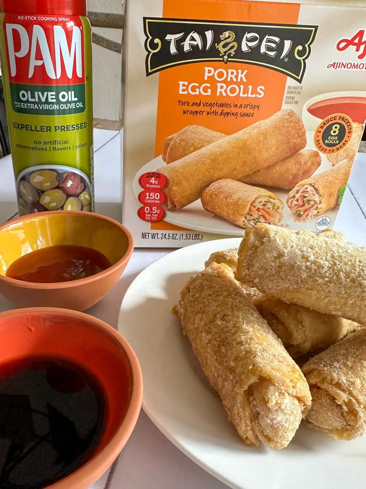 A package of Tai Pei Pork Egg Rolls, bottle of Pam Olive Oil Cooking Spray, frozen egg rolls on a white plate and 2 small bowls with dipping sauces.