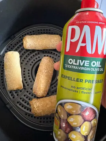 Egg rolls in an air fryer basket. There is someone holding a bottle of Pam Olive Oil Cooking Spray in the foreground.