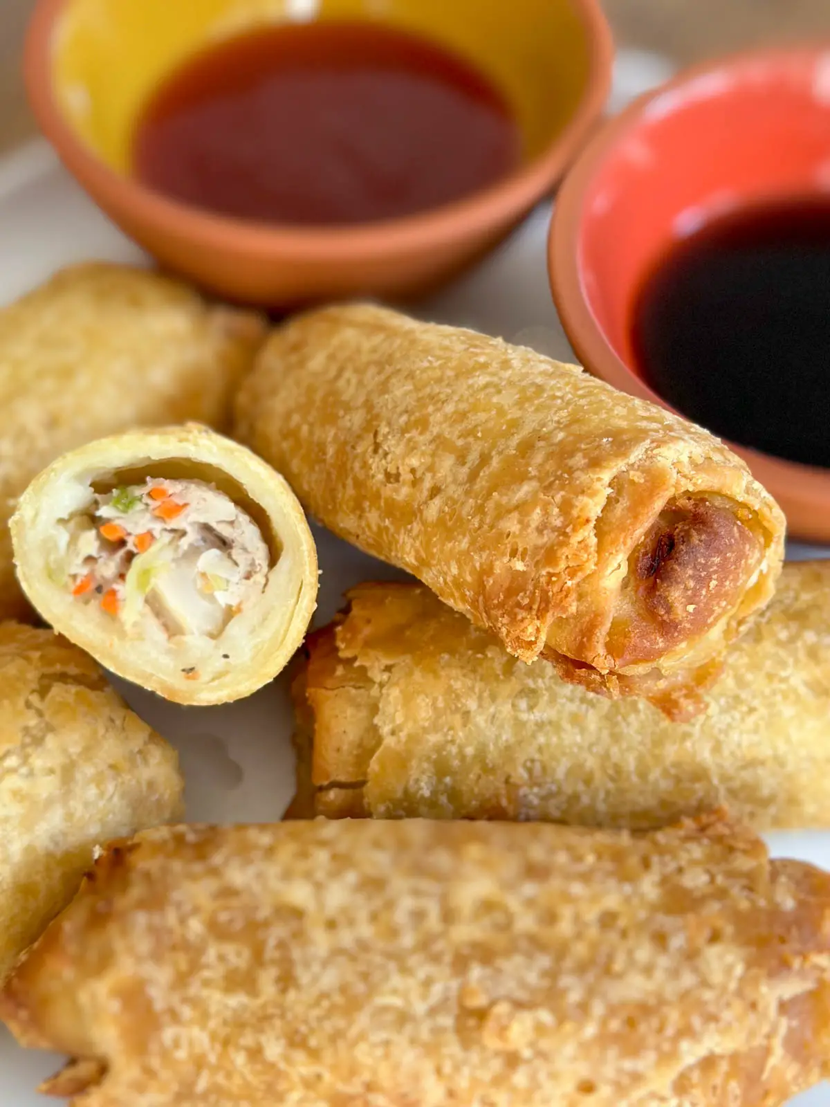 Cooked Egg rolls on a white plate. One is cut in half and there are 2 bowls with dipping sauces.
