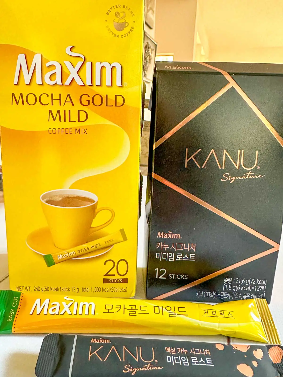 A package of Maxim Mocha Gold Mild Coffee Mix and a package of Kanu Signature coffee mix. There are individual sticks of each of these coffees in front of the packages.