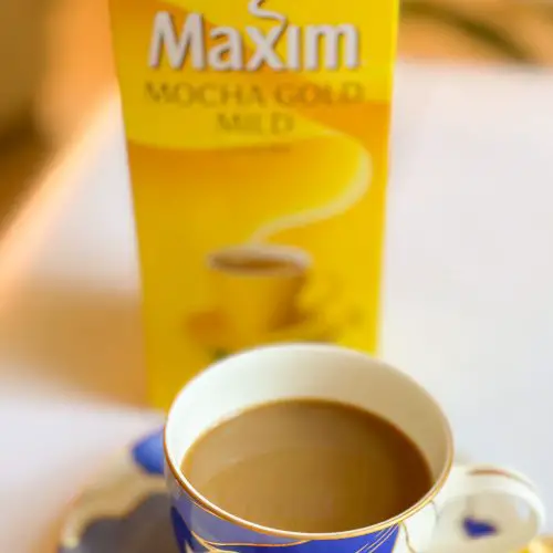 A package of Maxim Mocha Gold Mild in the background and a blue patterned coffee cup with coffee in the foreground with one of the Maxim coffee sticks on the plate the cup is resting on.