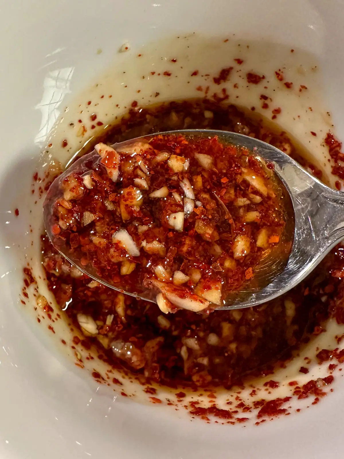 A white bowl containing a spicy sauce with Korean red pepper flakes, and garlic. There is a spoon containing some of the sauce in the foreground.