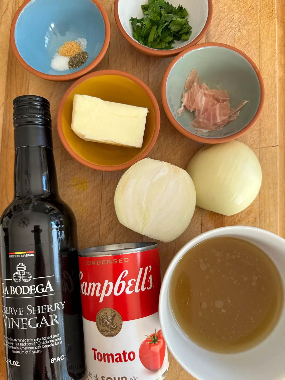 A can of Campbell's Tomato soup, a white bowl with chicken broth, a bottle of sherry vinegar, 2 onion halves, and small bowls containing butter, seasonings, parsley, and prosciutto.