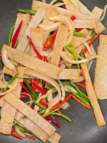 Sliced fish cake sheets with sliced onion and slivers of green and red peppers in a wok.