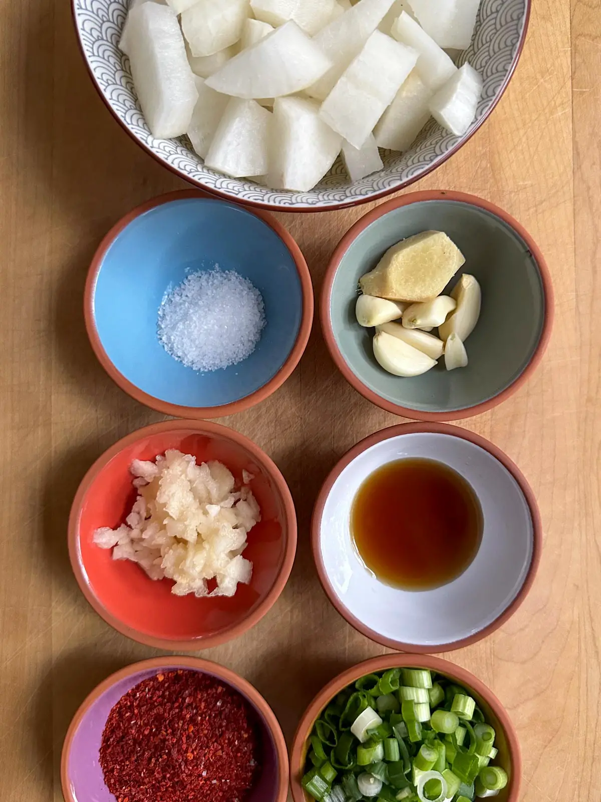 A bowl containing cubed pieces of daikon radish, and small bowls containing salt, ginger and garlic, Asian pear, fish sauce, red pepper powder, and green onions.