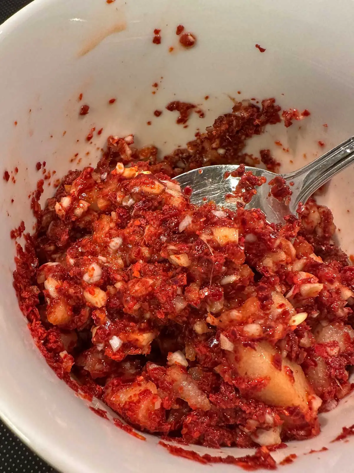 A white bowl containing a spicy red paste with red pepper powder, minced garlic, onion, and Asian pear. There is a spoon in the bowl containing some of the spicy paste.