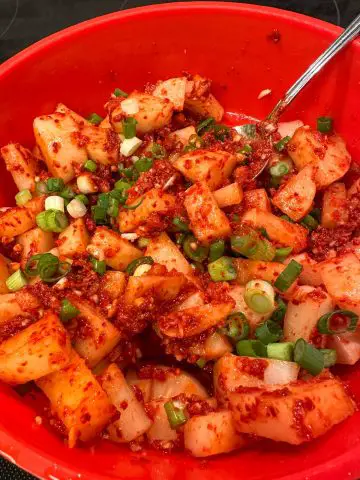 A red bowl containing radish kimchi which is cubed radish pieces in a spicy red sauce and green onions. There is a silver spoon in the bowl.