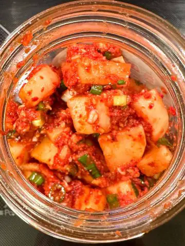 Daikon radish pieces in a spicy red sauce with green onions in a glass jar.