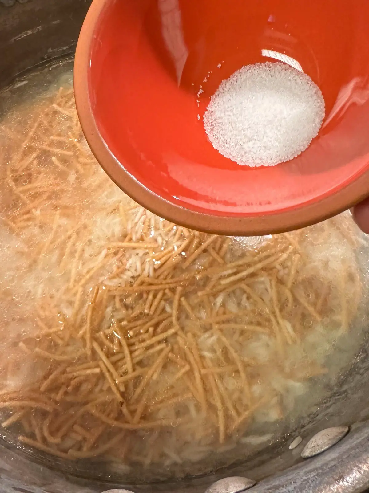 Rice and vermicelli in water in a saucepan with a small red bowl containing salt poised over the saucepan.