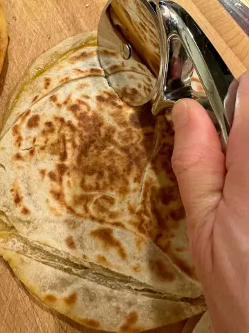 A browned quesadilla with a pizza cutter on top held by someone's hand demonstrating how to use it to cut the quesadilla tortilla into wedges.