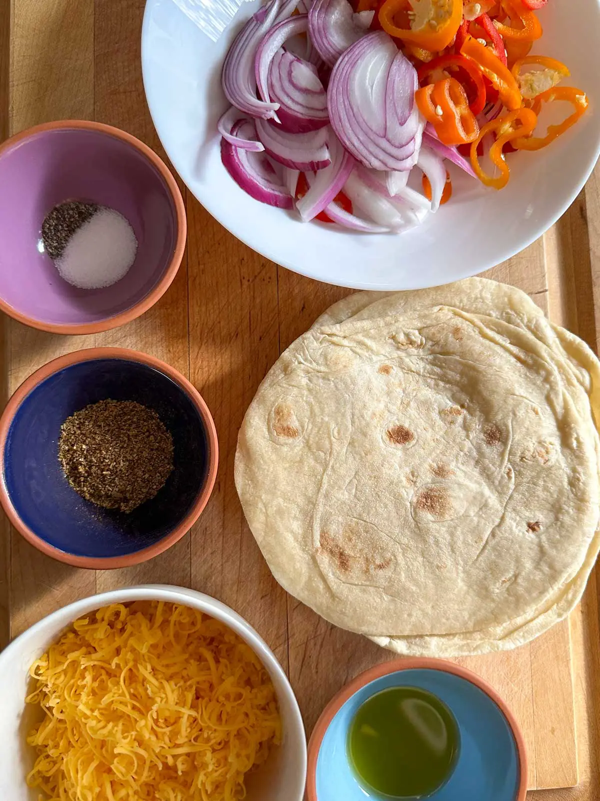 A white bowl containing sliced mini sweet peppers and sliced red onion, tortillas, and bowls containing shredded cheese, salt and pepper, fajita seasoning, and olive oil.