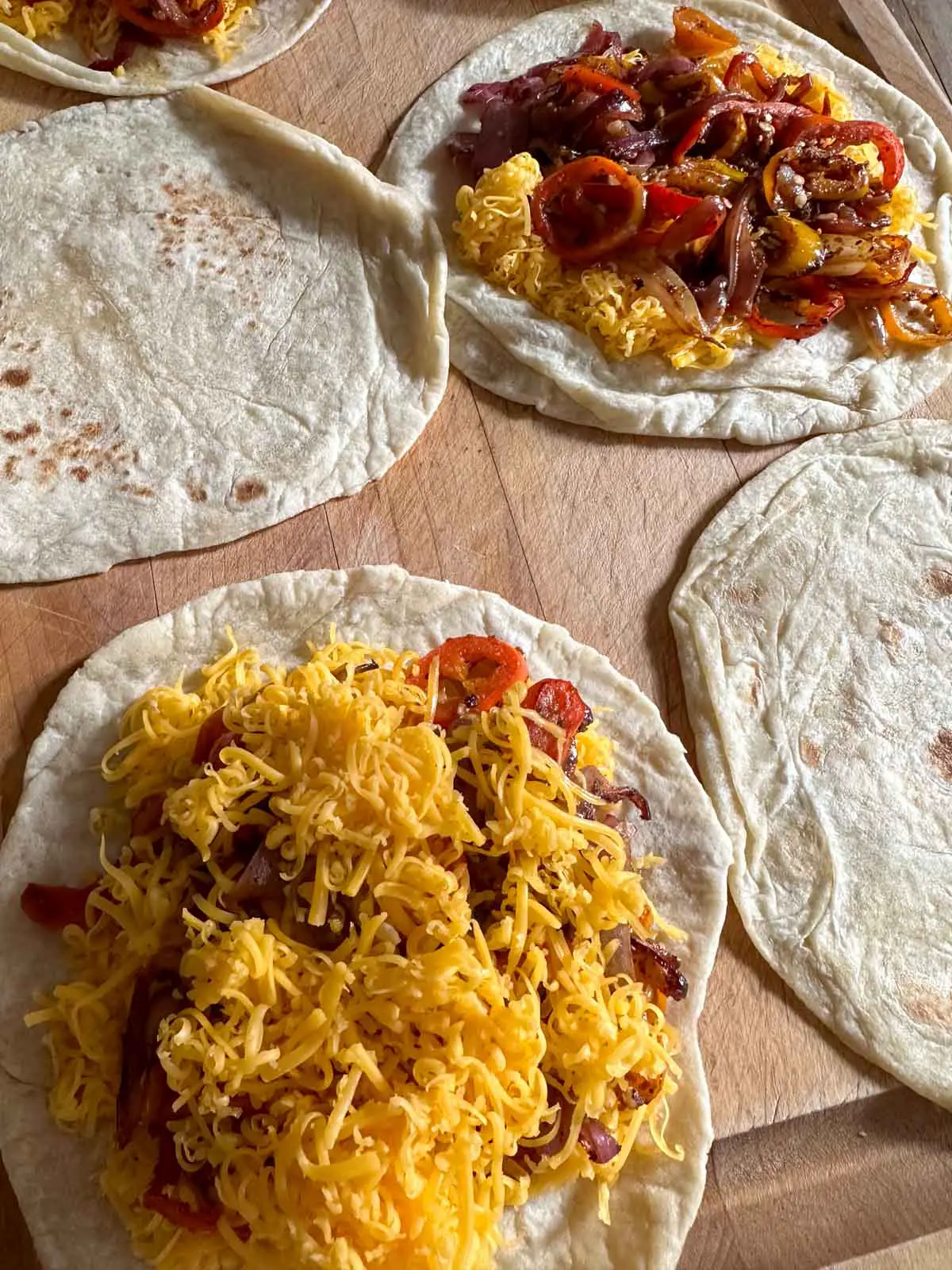 Tortillas placed on a wooden cutting board. 2 of the tortillas are topped with shredded cheese and a sautéed vegetable mixture.