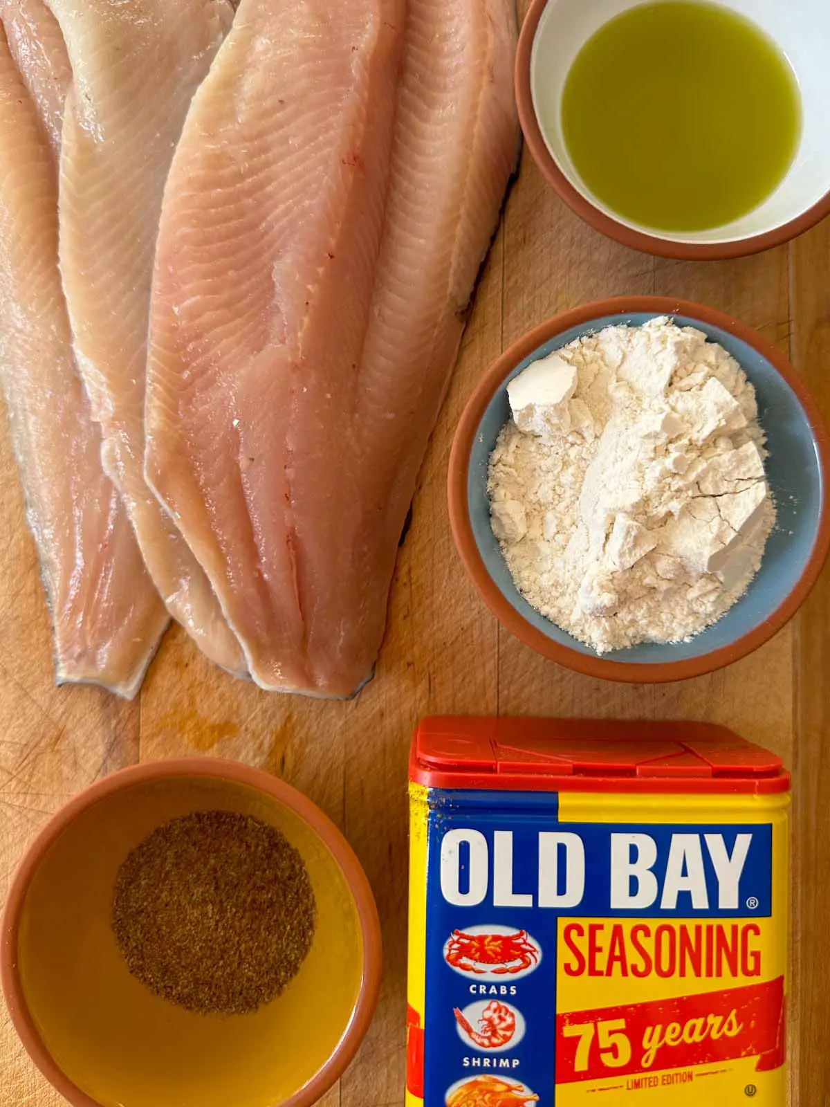 Rainbow trout fillets, bowls filled with olive oil, flour, and Old Bay Seasoning, and a container of Old Bay Seasoning.