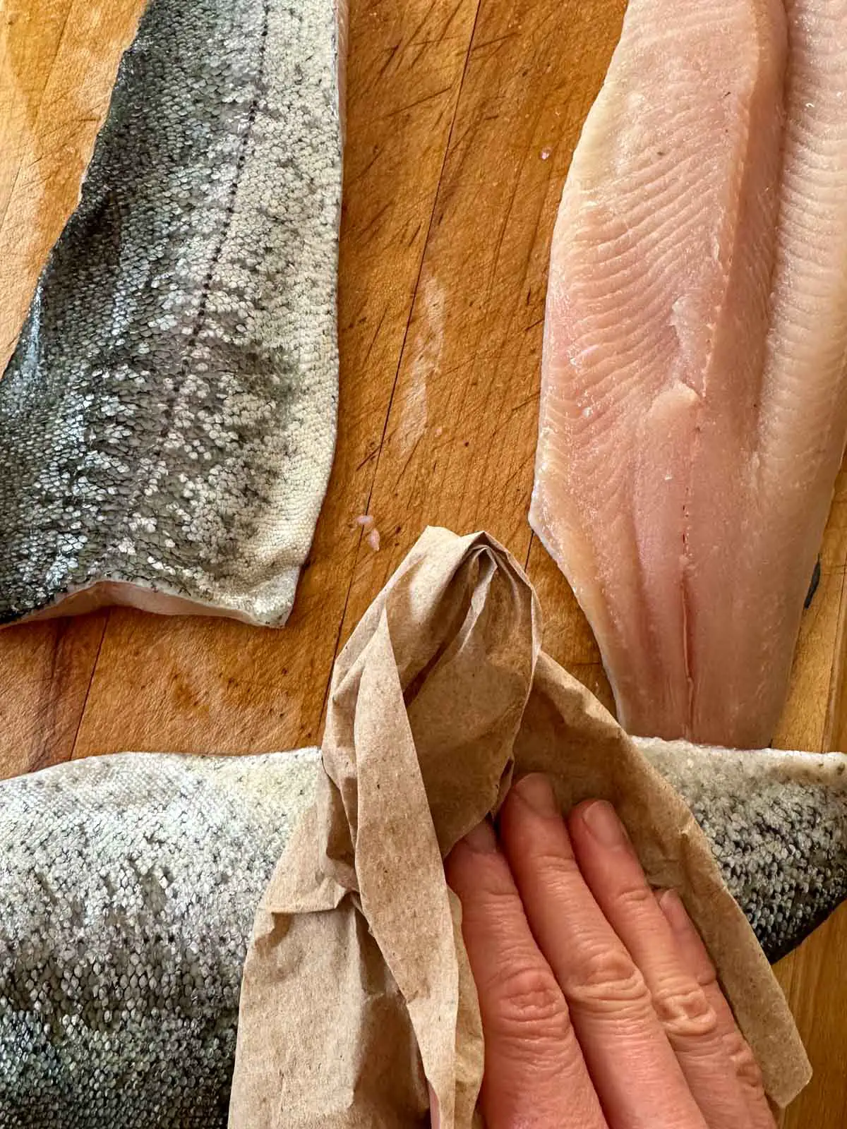 Rainbow trout fillets on a wooden cutting board with a paper towel being pressed down by a hand on one of the fillets to dry the fish.