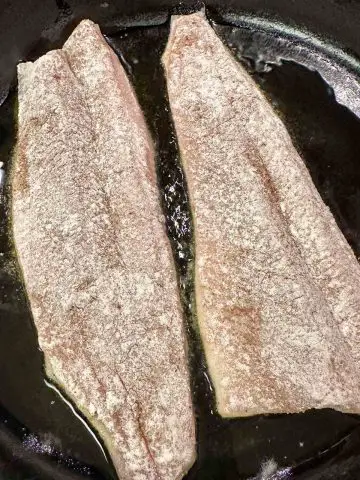 Rainbow trout fillets dredged in flour cooking in oil in a cast iron skillet.