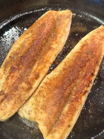 Seasoned rainbow trout fillets cooking in oil in a cast iron skillet.