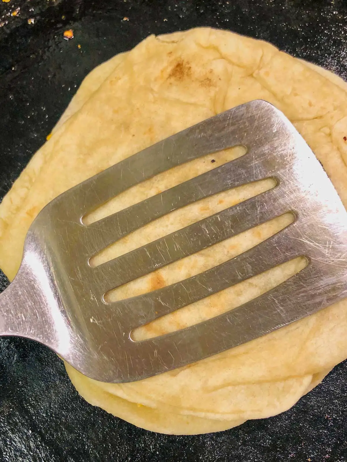 A metal spatula pressed down on a flour tortilla in a cast iron pan.