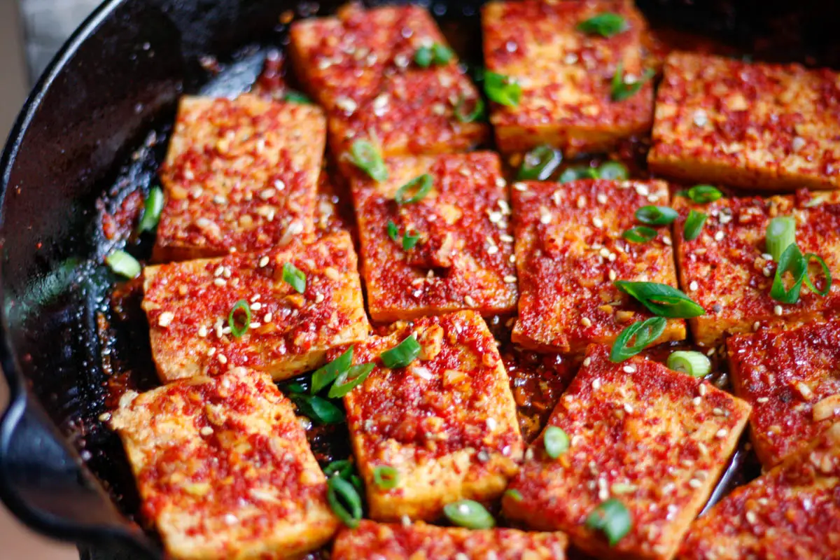 Tofu slices in a cast iron pan coated in a spicy sauce with sesame seeds and green onions as garnish.