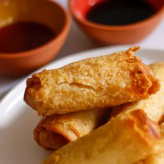 Crispy cooked egg rolls on a white plate with 2 small bowls containing dipping sauces in the background.
