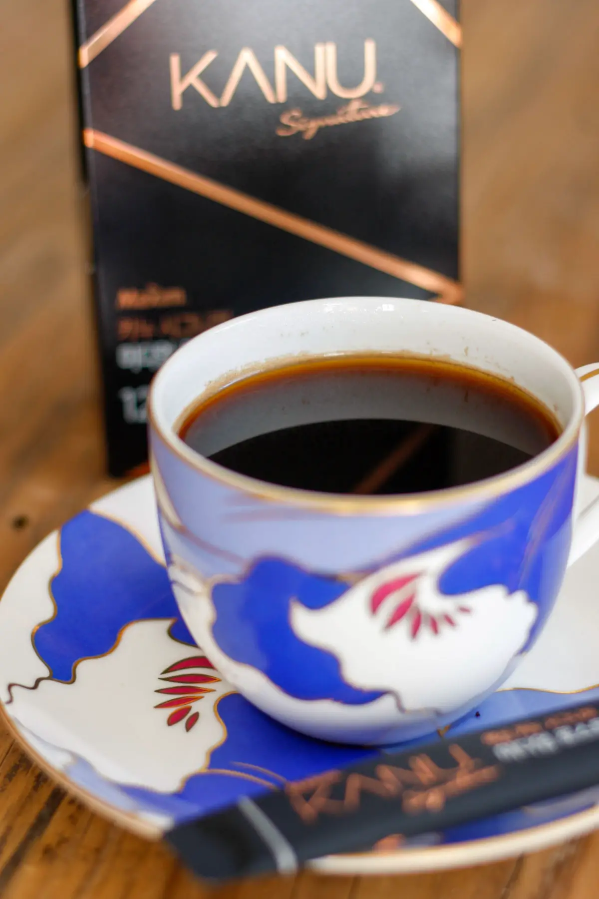 A package of Kanu Signature instant coffee in the background and and a blue patterned coffee cup with black coffee in the foreground with one of the Kanu signature coffee sticks on the plate the cup is resting on.