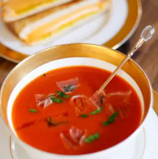 A gold rimmed bowl containing tomato soup garnished with parsley and prosciutto. There is a gold spoon with a glass top in the bowl. In the background there is grilled cheese sandwiches on a gold rimmed plate.