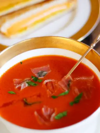 A gold rimmed bowl containing tomato soup garnished with parsley and prosciutto. There is a gold spoon with a glass top in the bowl. In the background there is grilled cheese sandwiches on a gold rimmed plate.