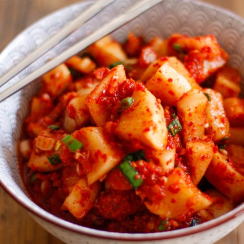 A patterned bowl containing cubed radish kimchi in a spicy red sauce with green onions. There is a pair of chopsticks resting on the upper left side of the bowl.