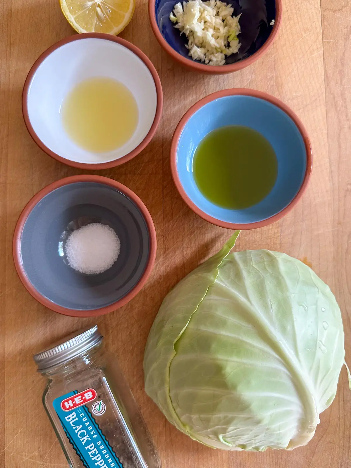 Green cabbage, jar labeled black pepper, bowls filled with salt, olive oil, lemon juice and garlic, and half a lemon.
