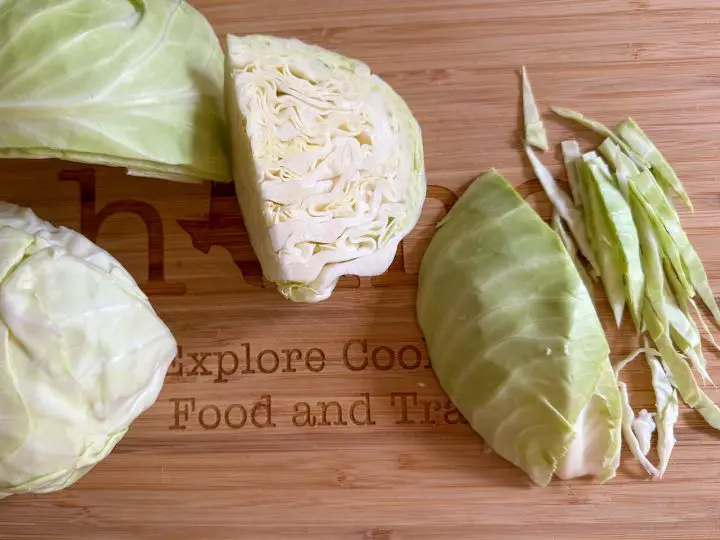 Green cabbage split into the white and green parts. Some of the cabbage has been cut into long green slivers. The cabbage is on top of a wooden cutting board.