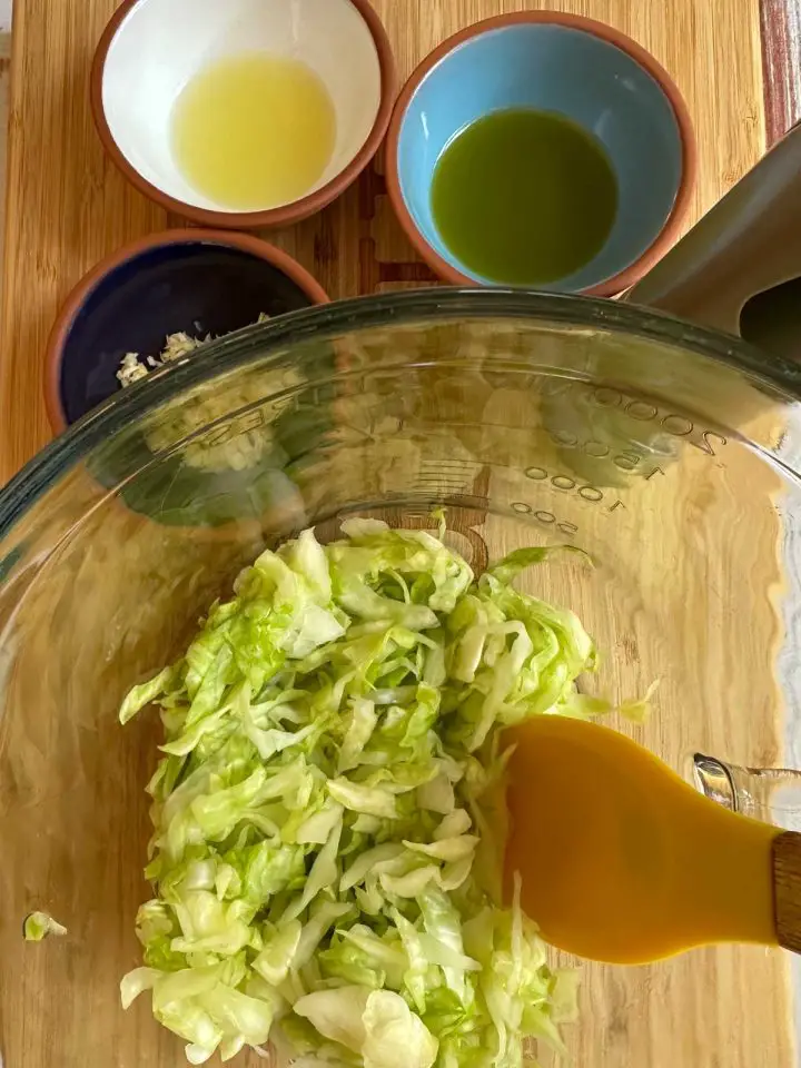 Shredded green cabbage in a glass bowl with a yellow spoon. There are small bowls containing lemon juice, olive oil, and garlic. There is a silver pepper grinder as well.