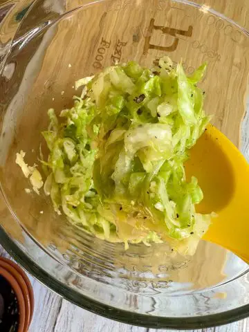 Green cabbage shredded to make a salad in a glass bowl, with a yellow spoon containing some of the cabbage salad. Black pepper has been added to the cabbage.