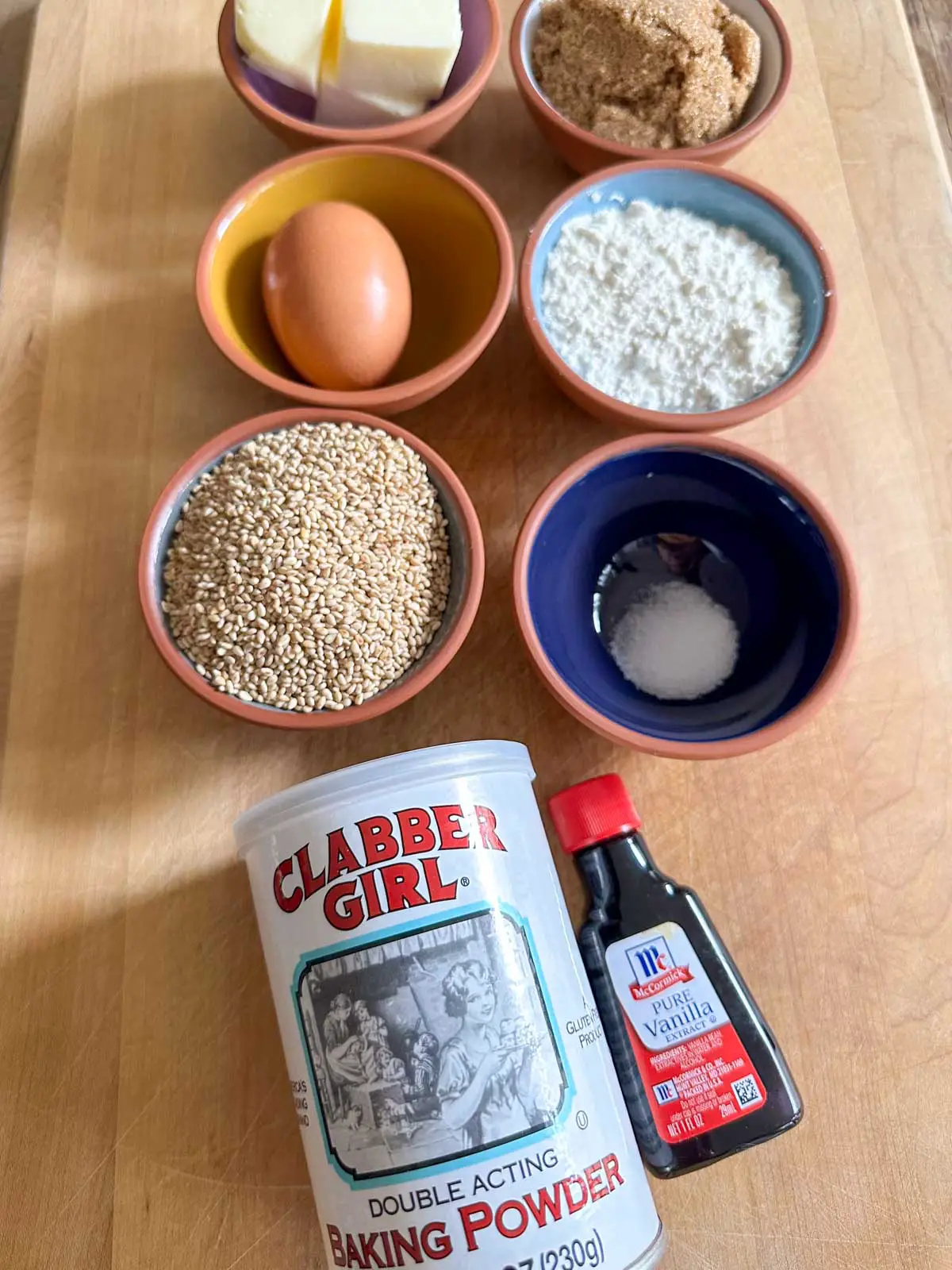 Small bowls containing butter, light brown sugar, an egg, flour, sesame seeds, salt, a can of Clabber Girl Baking Powder, and a small bottle of Vanilla Extract.