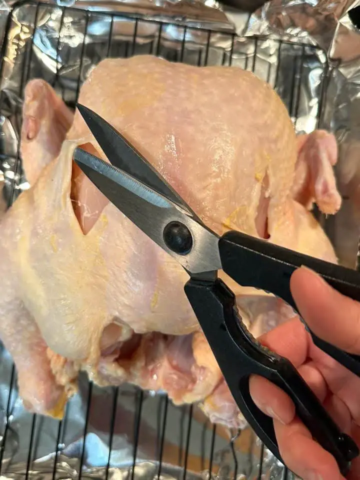 An uncooked whole chicken on a wire rack in a roasting tray lined with foil. There is a hand holding a pair of kitchen shears demonstrating cutting a whole in the skin of the chicken.