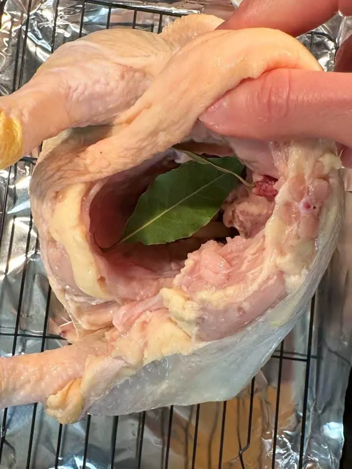 An uncooked whole chicken on a wire rack in a roasting tray lined with foil. There is a hand holding the chicken to show the cavity which contains bay leaves.