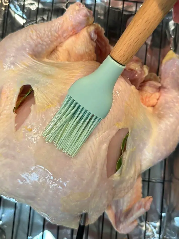 An uncooked whole chicken on a wire rack in a roasting tray lined with foil. There is a blue silicone baster with wooden handle basting the chicken with butter.
