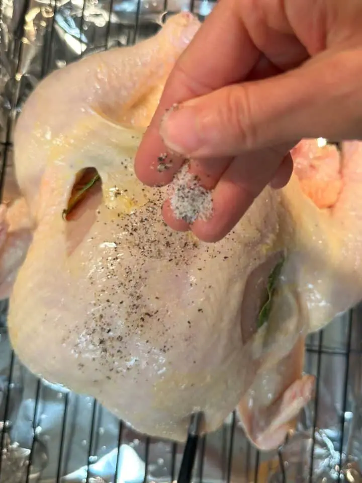 An uncooked whole chicken on a wire rack in a roasting tray lined with foil. There is a hand poised over the chicken sprinkling salt and pepper and a MEATER thermometer inserted into the chicken.