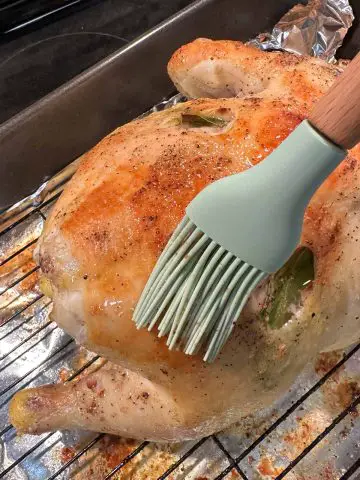 A whole chicken on a wire rack in a roasting tray lined with foil. There is a blue silicone baster with wooden handle basting the chicken with butter.
