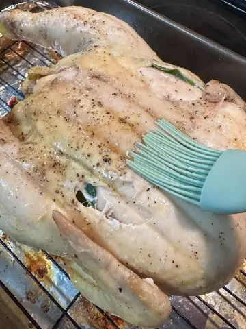 An uncooked whole chicken on a wire rack in a roasting tray lined with foil. There is a blue silicone baster with wooden handle basting the chicken with butter.