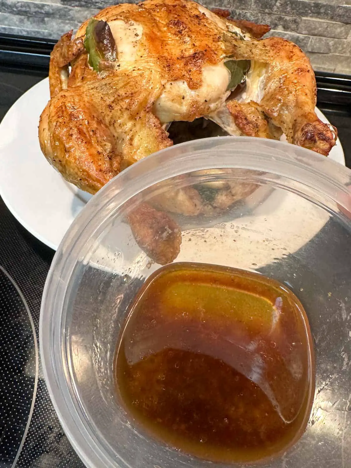 A roast chicken with crispy skin stuffed with garlic and bay leaves on a white plate. There is a plastic container with chicken fat drippings in the foreground.