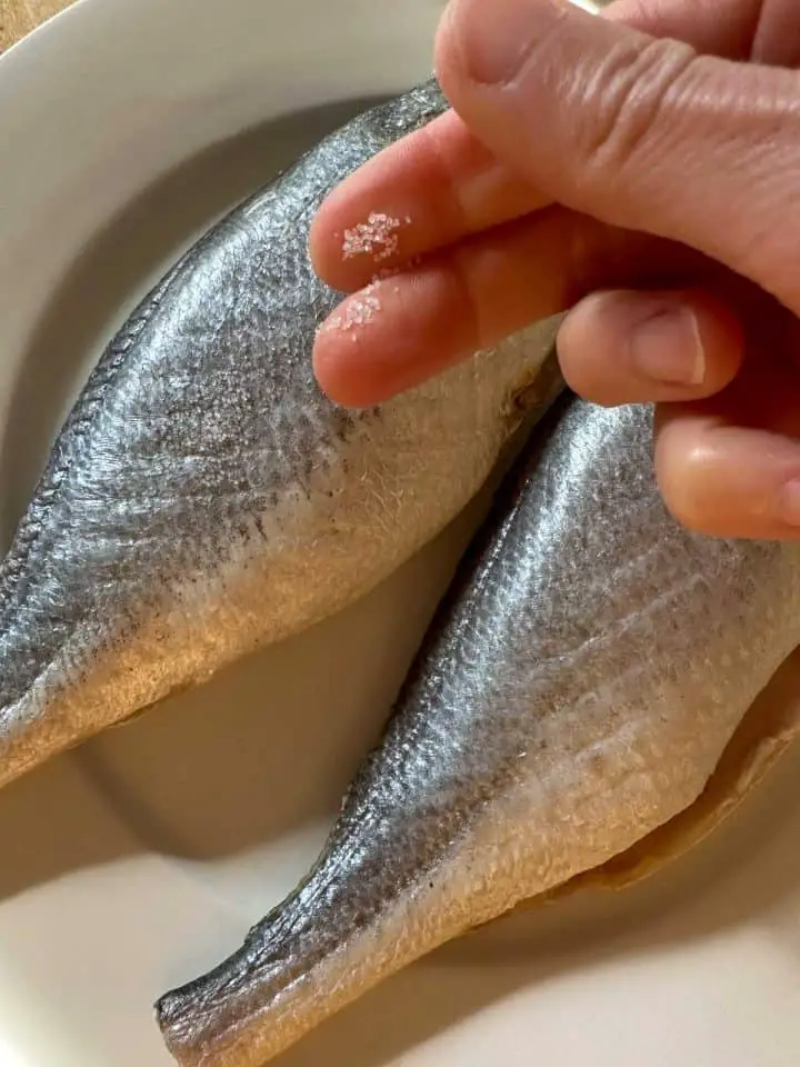 Two corvina fish on a white plate. There is a hand poised over the fish which has been sprinkled with salt and salt on some of the fingers on the hand.