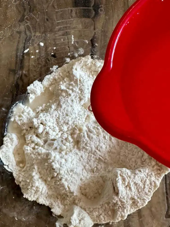 A glass bowl containing flour with a red measuring cup containing water poised above the flour.