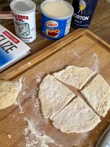A cutting board with uncooked dough cut into triangular shapes and a dough ball. There are containers of salt, Crisco, and baking powder on the side and a Belize travel book.