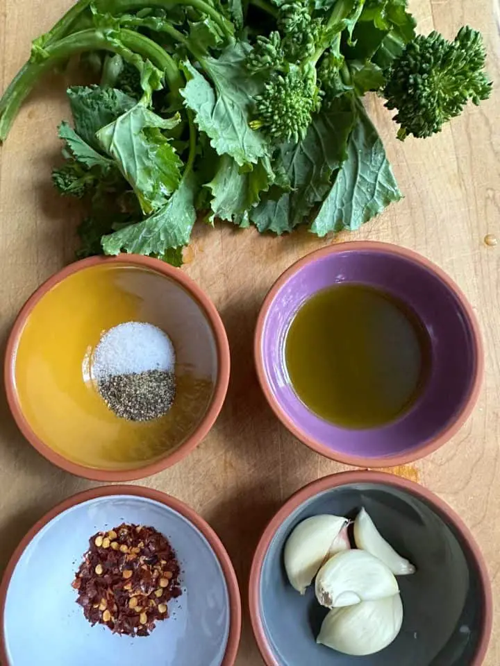 A bunch of rapini on a wooden cutting board along with small bowls containing salt and pepper, olive oil, chili flakes, and garlic cloves.
